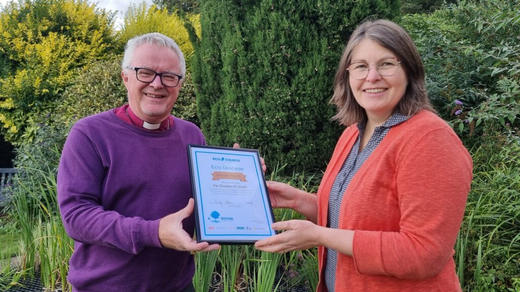 Bishop David Court handing the A Rocha UK Eco Diocese award  to Canon Sarah Spencer, Mission and Discipleship Officer and Envrionmental Officer for the Diocese. They are smiling. There is lots of green nature in the background. 