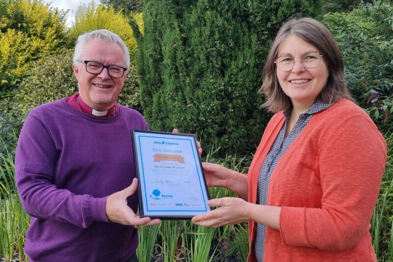 Bishop David Court handing the A Rocha UK Eco Diocese award to Canon Sarah Spencer, Mission and Discipleship Officer and Envrionmental Officer for the Diocese. They are smiling. There is lots of green nature in the background.